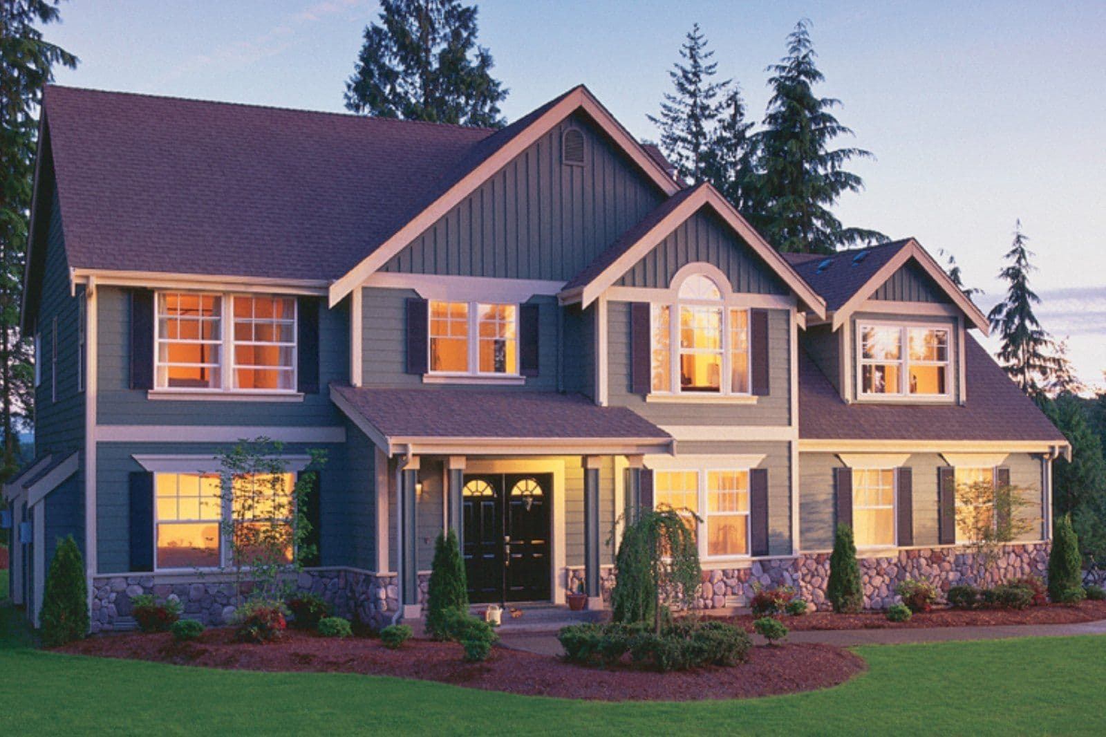 A blue house with light-colored trim, black double front door and black shutters at dusk.